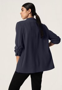 Blusa azul marino en tela ligera con mangas remangadas y ajuste holgado, con cuello y detalle de canesú en la espalda. Combinada con pantalones negros.