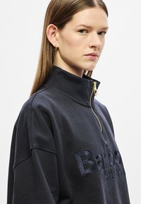 Μαύρο φούτερ με ψηλό γιακά, κλείσιμο με φερμουάρ έως το ένα τέταρτο και κεντημένο λογότυπο στο μπροστινό μέρος. Ομαλή υφή και άνετη εφαρμογή.