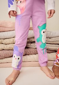 Lila barnbyxor med stora ugglegrafik i grön och lila, med ribbade muddar vid anklarna. Mjuk, bomullsliknande textur.