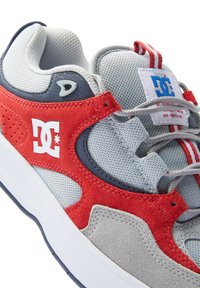 DC boty, s šedou síťovinou a červenými semišovými akcenty. Logo na boku, polstrovaný jazyk a šedé tkaničky. Výrazný červený a bílý pruhovaný detail.