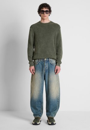 Jonge man met een donkergroene gebreide trui, losse vervaagde blauwe jeans, groene en beige sneakers en grote donkere zonnebril, staand tegen een effen achtergrond.