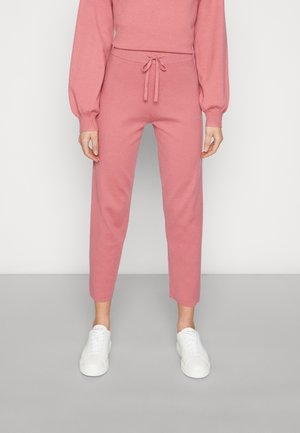 Persoon die hoog getailleerde roze gebreide broek met trekkoord en witte sneakers draagt, staand tegen een lichtgrijze achtergrond.