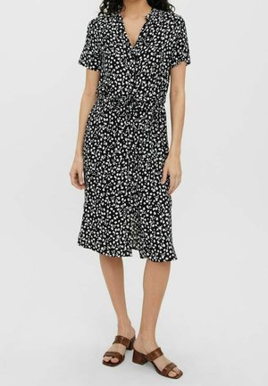 Žena v černých midi šatech s bílým květinovým potiskem, krátkými rukávy a hnědými sandály na podpatku, stojící před jednoduchým světlým pozadím.