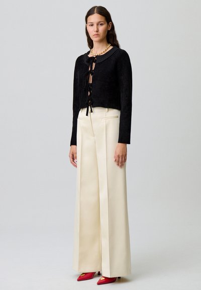 Pull noir à texture, court avec des liens à l'avant, associé à un pantalon large crème à taille haute et des chaussures rouges à bout pointu.