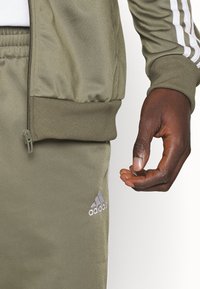 Olivovo zelený Adidas teplákový súpravy so zipsovými vreckami. Obsahuje biele trojpruhové akcenty na rukávoch a logo na ľavom nohavici. Hladká textúra.