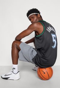Ténis de basquetebol preto com detalhes em verde, número 5, calções cinzentos, sapatilhas brancas, sentado numa bola de basquetebol, fundo branco.