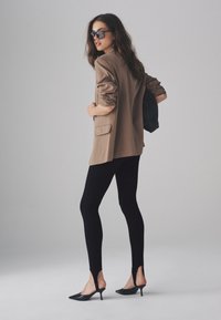 Pruun kohandatud bleiser, mustad retuusid lõhikuga ja mustad terava ninaga kingad. Modell kannab suurendatud päikeseprille ja musta kotti.