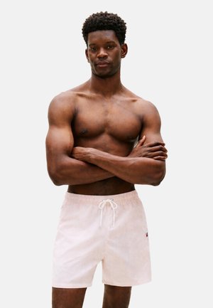 Hombre musculoso sin camiseta con los brazos cruzados que lleva puestos pantalones cortos de baño rosa claro con cordón, sobre un fondo liso.