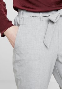 Pantalon taillé gris clair en tissu texturé, doté d'une ceinture à nouer et de poches latérales. Le vêtement est associé à un haut bordeaux.