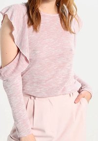 Haut rose clair avec volants aux épaules et manches longues, fabriqué dans un matériau doux et texturé. Présente une coupe décontractée et un col arrondi.