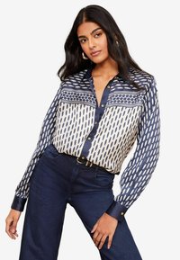 Navy-blaues und cremefarbenes gemustertes Bluse mit Knopfleiste, die geometrische Designs und lange Ärmel aufweist. Kombiniert mit dunkelblauen Jeans.