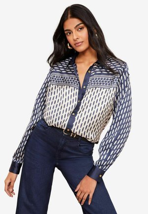 Blusa a righe blu navy e crema con bottoni davanti, caratterizzata da design geometrici e maniche lunghe. Abbinata a jeans blu scuro.
