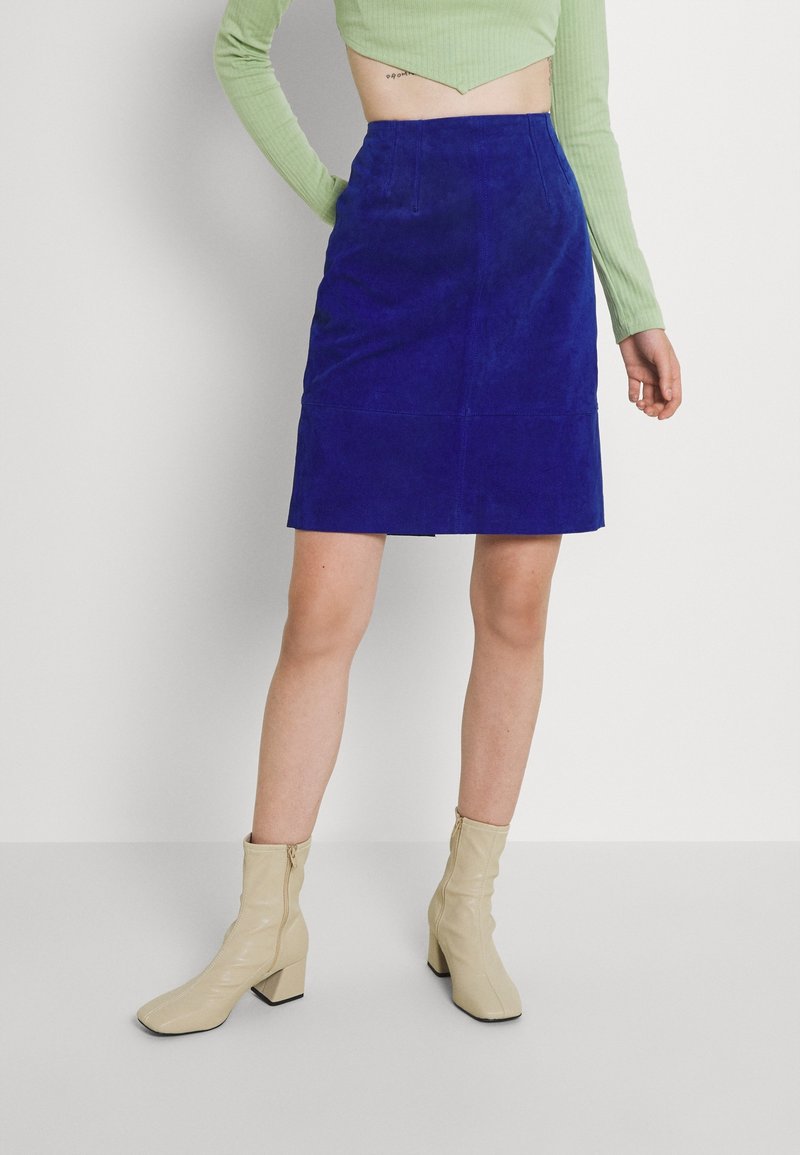 Jupe en daim bleu à coupe ajustée, longueur au genou, avec des coutures décoratives. Associée à des bottines beiges et un haut vert côtelé.