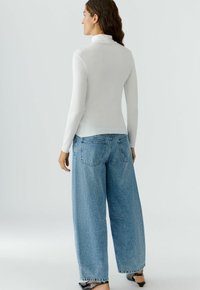 Weißes Langarm-Turtleneck kombiniert mit hellblauen weit geschnittenen Jeans. Das Oberteil hat eine glatte Textur; die Jeans sind mit Gesäßtaschen ausgestattet und haben eine lockere Passform.