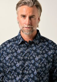 Mittelalter Mann mit grauem Haar und Bart, der ein navyblaues Shirt mit blau-weißen Blumenmustern trägt, blickt gegen einen hellen Hintergrund.