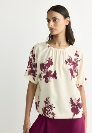 Woman wearing cream blouse with burgundy floral patterns and matching burgundy skirt, standing against plain light background.