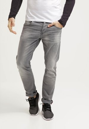 Mand iført grå jeans, en hvid skjorte med sorte ærmer og sorte sneakers; den ene hånd i lommen, står på en ensfarvet baggrund.