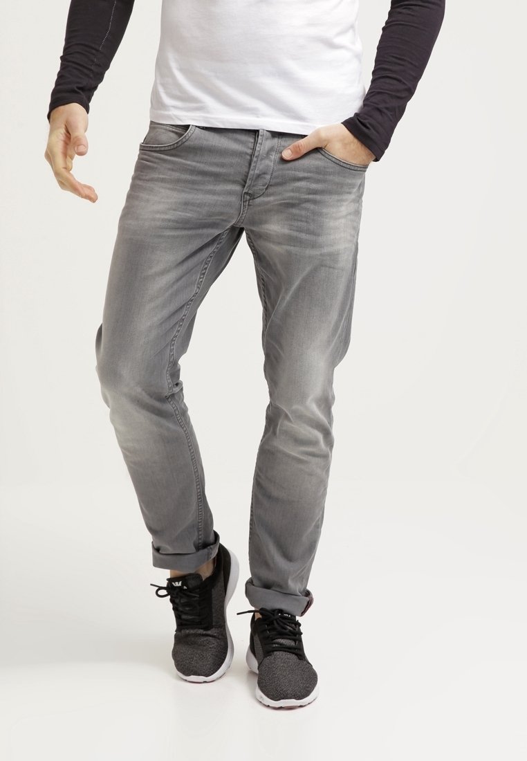 Homme portant un jean gris, une chemise blanche à manches noires et des baskets noires ; une main dans la poche, debout sur un fond uni.
