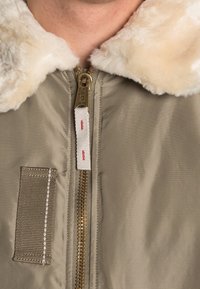 Giacca bomber beige con colletto in pelliccia sintetica, zip frontale e dettagli con tasche verticali. Tessuto morbido con finitura liscia e lucida.