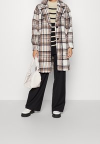 Manteau en laine à carreaux blanc, marron et noir, avec un large col, une fermeture à boutons et une coupe décontractée, associé à un pantalon noir et des bottes blanches.