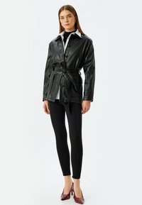 Giacca nera in ecopelle con chiusura a bottoni, vita cintata e colletto bianco, abbinata a leggings neri e tacchi rossi lucidi.