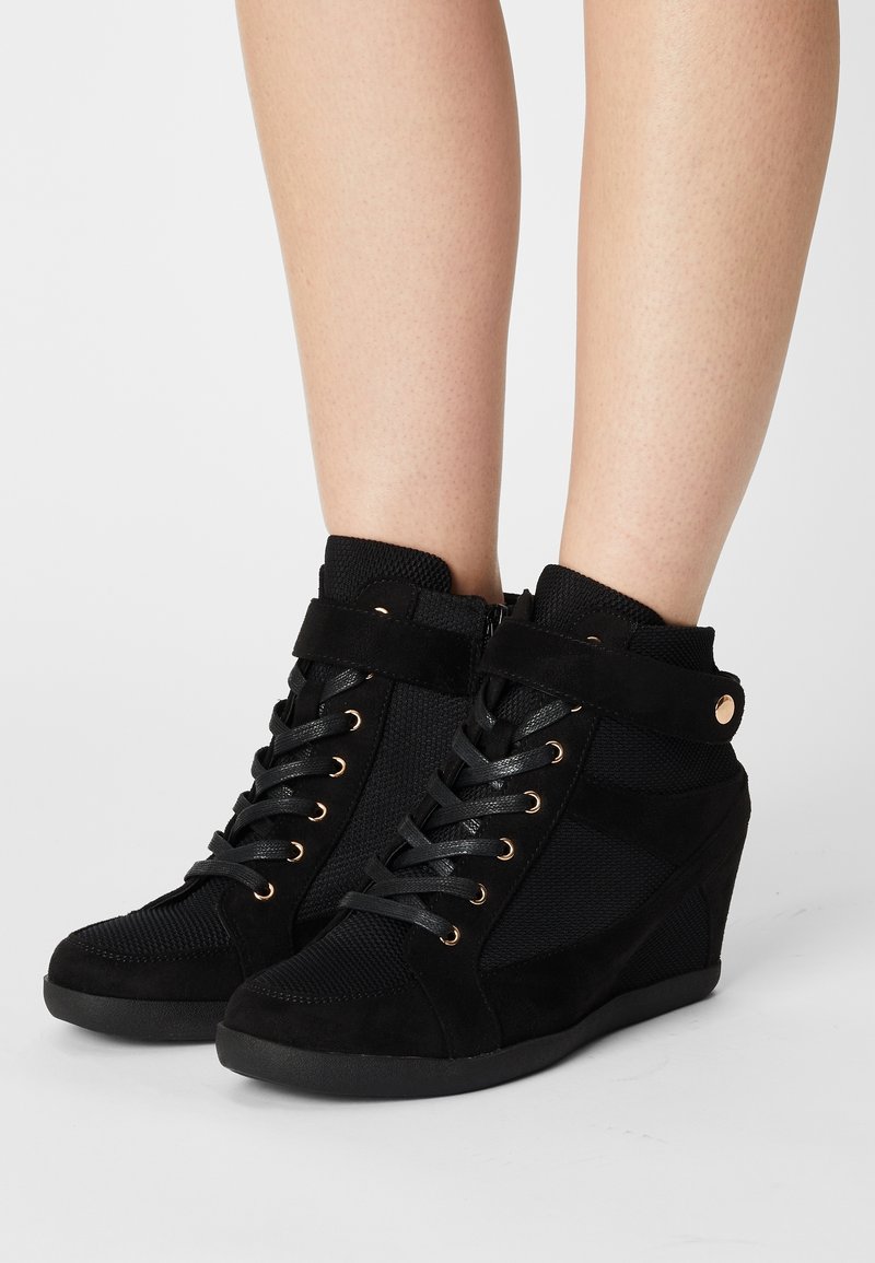 Svarta kilskor i mocka och mesh, med guldfärgade snören och detaljer, axelhöjd och strukturerade ytor.