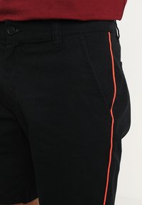 Černé šortky vyrobené z bavlny s úzkým střihem, s výrazným oranžovým bočním pruhem a standardními kapsami. Nastavitelný pás s knoflíkovým zapínáním.