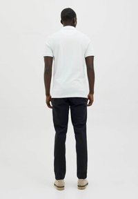 Witte polo met een kraag, gecombineerd met donkerblauwe broek. Model draagt lichte schoenen en staat tegen een plain witte achtergrond.