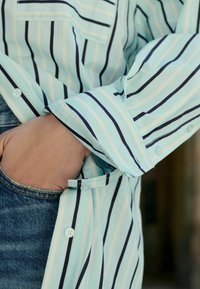 Lyseblå skjorte med tynde mørke striber, der har en brystlomme og knapdetaljer. Ærmet er oprullet, hvilket afslører denim nedenunder.