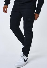 Pantalones cargo negros hechos de una tela texturizada, con bolsillos laterales y un detalle de botón. Llevados con zapatillas blancas.