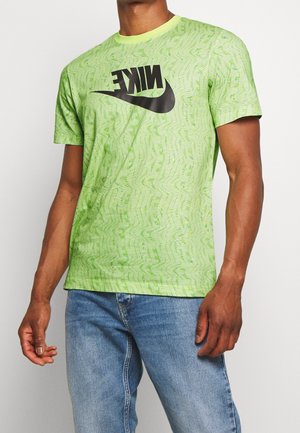 Mężczyzna w jasnzie zielonej koszulce Nike z wzorem, z czarnym odwróconym logo oraz niebieskich dżinsach, stojący na jednolitym tle.