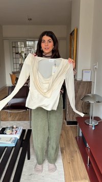 Langärmliges cremefarbenes Oberteil mit drapiertem Ausschnitt und Stoffraffungen, aus weichem Material, kombiniert mit olivgrünen Hosen. Etikett sichtbar.