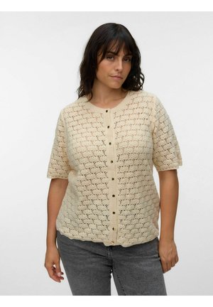 Kvinde med mørkt hår iført en cremefarvet, kortærmet hæklede cardigan med knapper foran og grå jeans mod en ensfarvet baggrund.