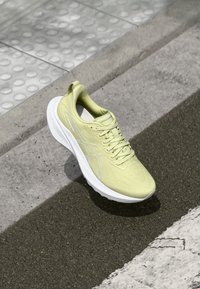 Chaussure de sport jaune clair avec une tige texturée, semelle blanche et lacets. Bout arrondi et languette au talon ; détail de marque minimal.