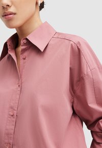 Camisa de botones de color rosa claro con un cuello estructurado, mangas largas y un corte relajado. Textura suave con detalles de botones visibles.