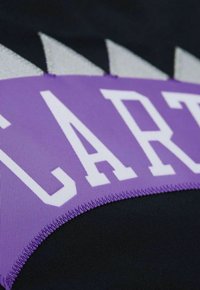 Tessuto nero con una fascia viola che presenta lettering bianco prominente e accenti argento frastagliati lungo il bordo superiore.