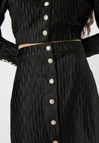 Sort to-delt sæt med en tekstureret, bølget design i skinnende stof. Det har metalknapper ned ad fronten af den cropped top og nederdelen.
