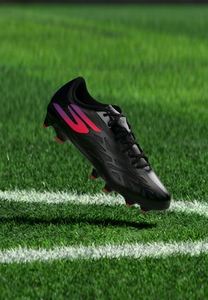 Crampon de football noir avec des rayures rouges et violettes flottant au-dessus de l'herbe verte près des lignes blanches du terrain.