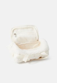 Hvit fleece-fôret bag med en glidelåslomme i mesh, myk tekstur, rund form og vevde stropper. Åpningen på toppen avslører innvendig plass.