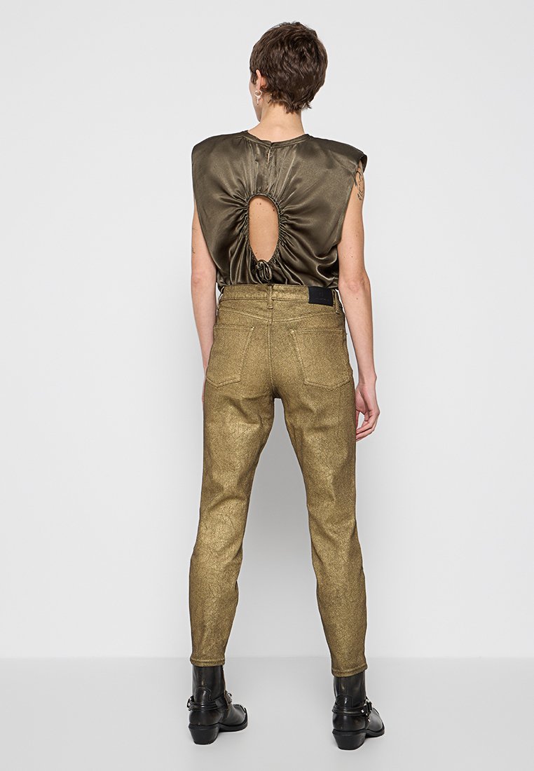 Oliwiana satynowa bluzka z tylnym rozcięciem i sznurkiem, w zestawieniu ze złotymi metalicznymi, dopasowanymi spodniami. Widoczne czarne botki za kostkę.