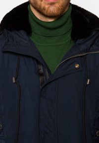 Granatowa parka z futrzanym kołnierzem, z zamkiem błyskawicznym i regulowanymi troczkami. Zielony sweter z golfem widoczny pod spodem.