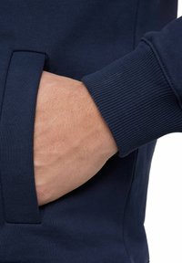 Felpa blu navy con polsini a costine, caratterizzata da una mano in una tasca laterale. Il tessuto sembra morbido con una finitura texture.