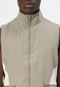 Sleeveless vest in light beige nylon with a high collar, front zipper, and white side accents. Smooth texture with minimal seams.