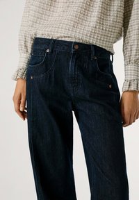 Jeans de mezclilla azul oscuro con un diseño de cintura alta y piernas anchas, y costuras visibles. Accesorios de hardware en tono cobre. Tejido texturizado.