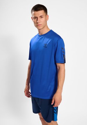 Jonge man in blauwe sportkleding met hummel-logo, staand tegen een effen witte achtergrond, recht vooruit kijkend met een neutrale uitdrukking.