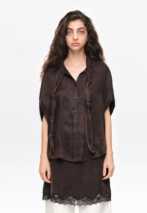Mujer con el pelo largo y rizado, vistiendo una blusa abotonada suelta de color marrón oscuro y una falda con ribete de encaje, de pie frente a un fondo blanco liso.