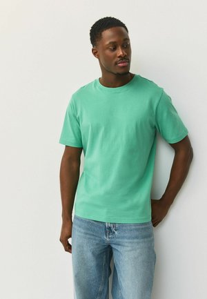 Młody mężczyzna w miętowo zielonej koszulce i jasnoniebieskich dżinsach stoi na tle białej ściany, lekko spoglądając na bok, z jedną ręką w kieszeni.