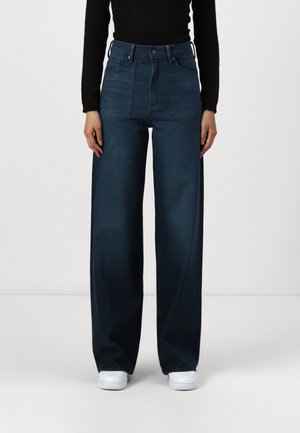 Kobieta ubrana w ciemnoniebieskie jeansy z wysokim stanem i szerokimi nogawkami, czarną bluzkę z długim rękawem oraz białe trampki, stojąca na jednolitym tle.