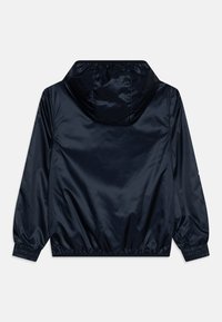 Casaco leve azul marinho com capuz, feito de um material brilhante e suave. Apresenta punhos e barra elásticos, design minimalista, sem fechos visíveis.
