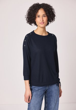 Donna con capelli ricci che indossa un maglione blu navy con bottoni sulle spalle e jeans blu, in piedi davanti a uno sfondo chiaro e uniforme.